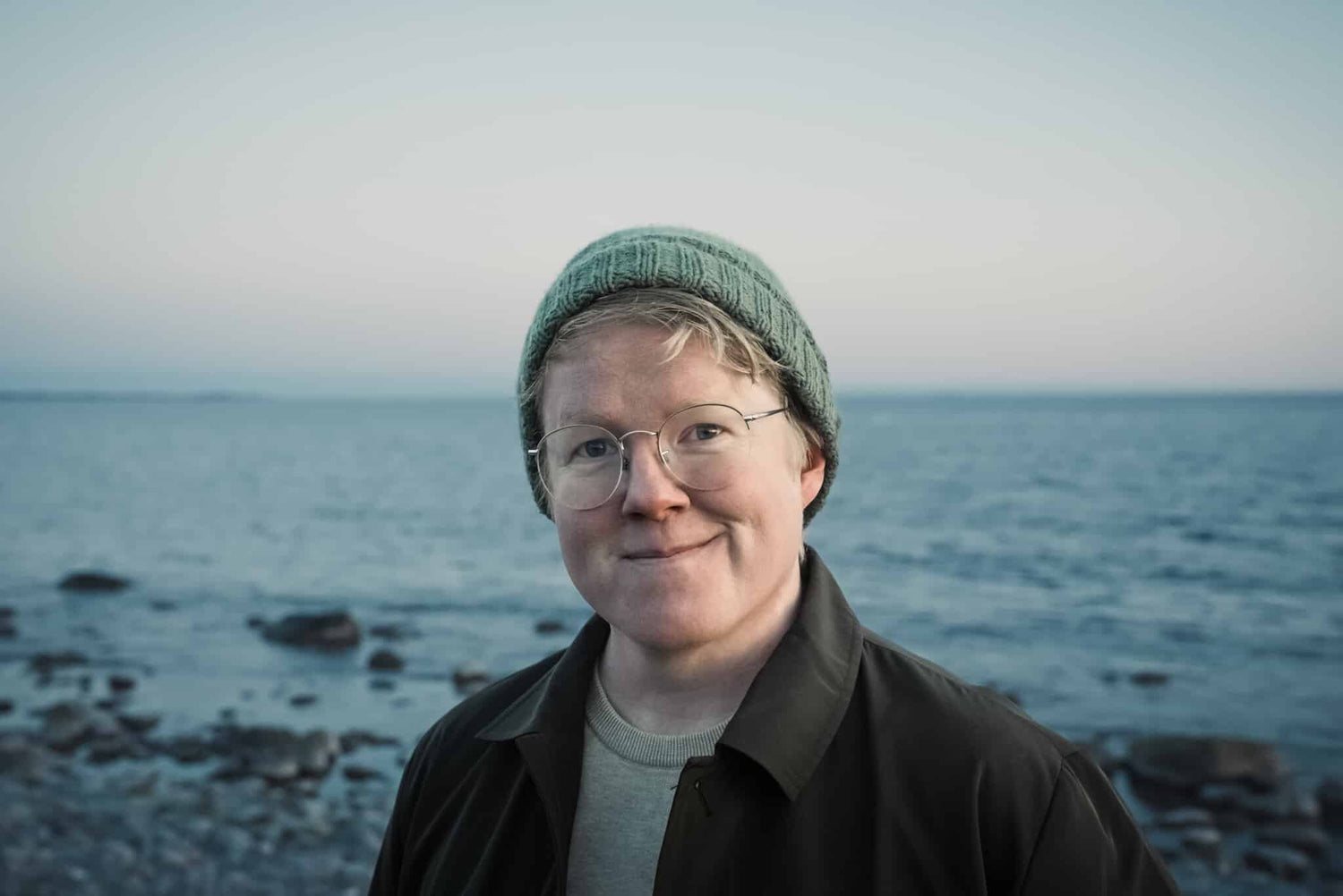 Person wearing a green beanie and glasses standing on a rocky beach with ocean in the background